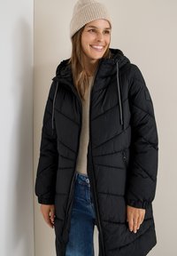 Sort puffjakke med lynlås, hætte og snore. Glat tekstur med chevron-syningsdetaljer. Båret over en beige trøje og blå jeans.
