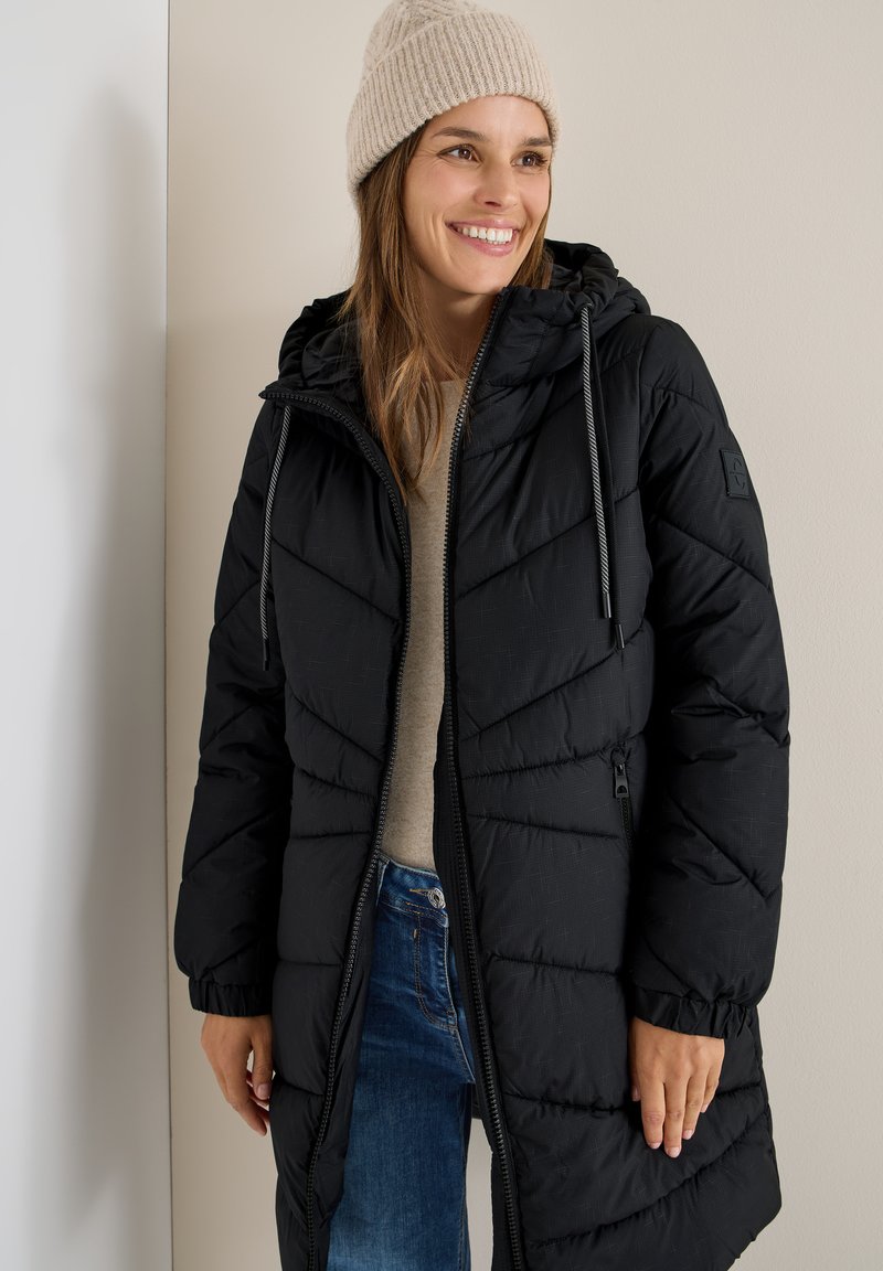 Chaqueta de plumas negra con cierre de cremallera, capucha y cordones. Textura suave con detalles de costura en forma de chevron. Llevada sobre un suéter beige y jeans azules.