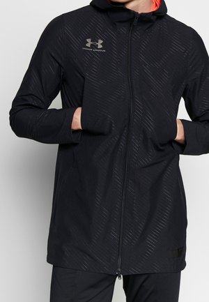 Czarna kurtka z kapturem Under Armour z subtelnymi ukośnymi paskami, z przednim zamkiem błyskawicznym i rękami w bocznych kieszeniach, noszona na pomarańskiej koszulce.