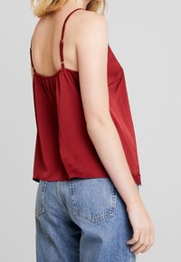 Rød satin cami med justerbare spaghetti stropper, en rynket ryg og blonde detaljer ved kanten, parret med lyse denimjeans.