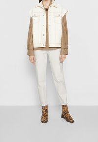 Chaleco de mezclilla blanco con tirantes en los hombros, suéter de punto texturizado marrón, pantalones beige claros y botines de puntada animal marrón con tacón de bloque.