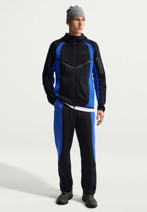 Ung mand iført en sort og blå lynlåstrøje, matchende bukser, grå hue og sorte sneakers, stående mod en ensfarvet lys baggrund.