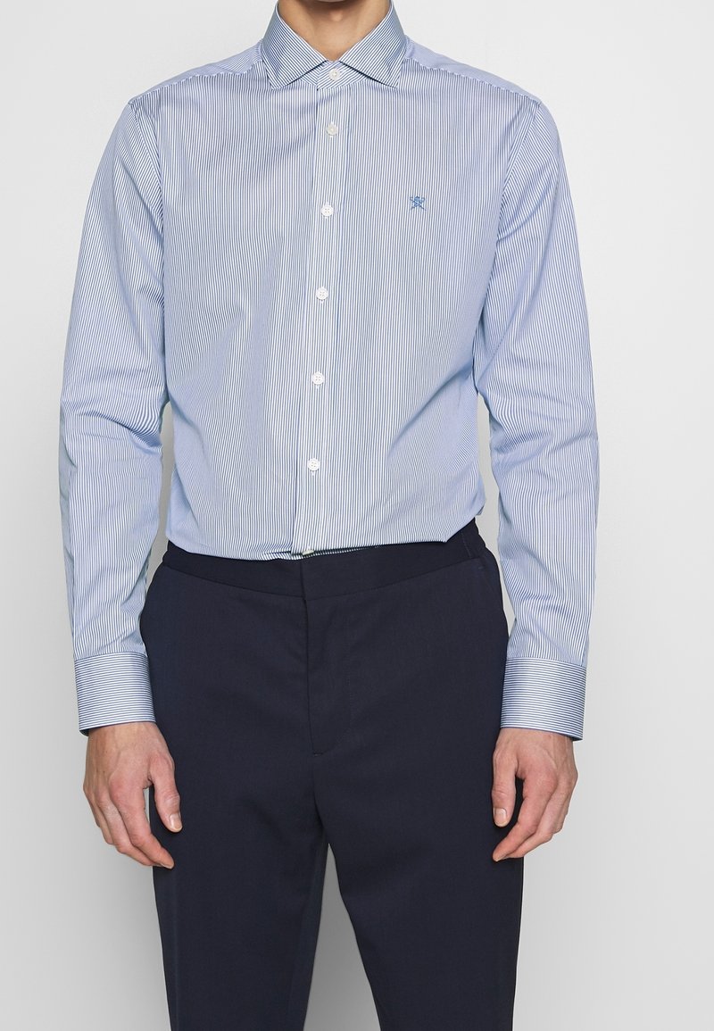 Homme portant une chemise boutonnée à manches longues à fines rayures bleues et blanches, rentrée dans un pantalon bleu marine foncé, mains le long du corps.