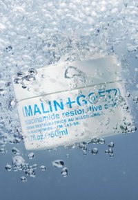 Niacinamid-reparaturcreme in einem weißen runden Glas mit blauer Schrift, im klaren Wasser eingetaucht, umgeben von Wassertropfen und Blasen.