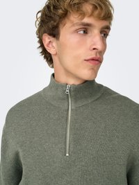 Pull vert côtelé avec un col montant et un détail de fermeture éclair argentée. Coupe ajustée et texture douce.