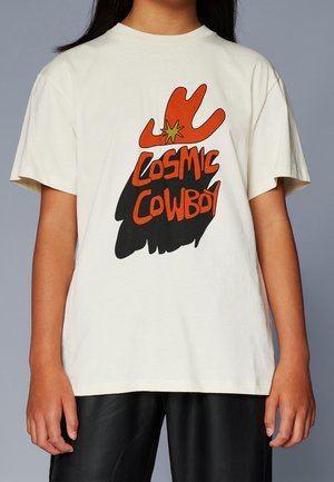 Person iført en off-white T-shirt med en rød cowboyhat grafik og ordene "Cosmic Cowboy" i rød og sort tekst.