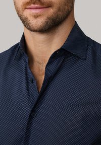 Man in een marineblauw knopshirt met witte stippen en een gedeeltelijk open kraag, zichtbaar stoppelbaard en borsthaar.
