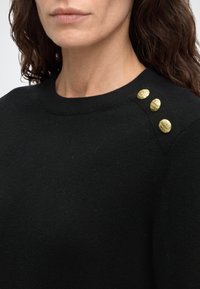 Femme portant un pull noir à col rond avec trois boutons dorés texturés sur l'épaule gauche.