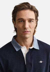 Marineblauw poloshirt met een lichtblauwe kraag, voorzien van een witte knopenlijst en een logo op de borst. De stof is glad met een geribbelde textuur.