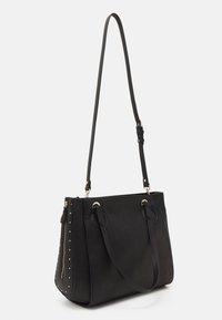 Bolso tote de cuero negro con tachuelas doradas en un lado, asas dobles y una correa para el hombro ajustable y removible. Textura suave.