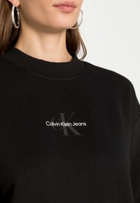 Sudadera negra con cuello acanalado y un logo frontal que incluye "Calvin Klein Jeans" en blanco con un sutil "CK" en gris.