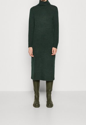 Femme portant une robe pull à col roulé vert foncé associée à des bottes hautes vert olive, debout devant un fond blanc uni.