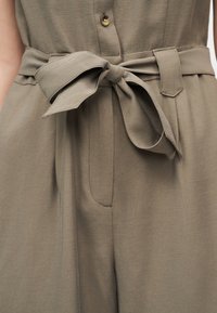 Κοντινή λήψη παντελονιού από ελαιοπράσινο ύφασμα με μπροστινό κουμπί, πιέτες και ζώνη μέσης δεμένη στη μέση.