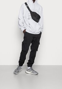 Grå hoodie med tryckt text, svarta cargo-byxor med fickor, och grå sneakers med vit sula. Axelväska i svart tyg.