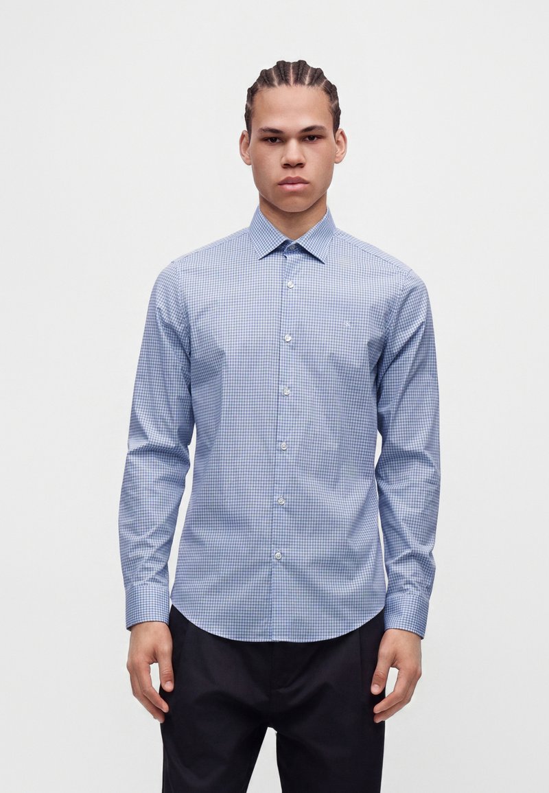Jeune homme aux cheveux tressés portant une chemise bleue à petits carreaux et un pantalon noir, debout devant un fond clair uni.