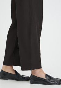 Mocasines negros de cuero con punta redondeada y detalle sutil de flecos, usados con pantalones negros de pierna ancha. Textura suave y diseño minimalista.