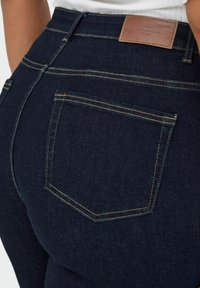 Tumši zilas džinsi ar vara diegiem, ar ādas zīmolu uz jostas vietas un aizmugurējo kabatu ar izliekto šuvju.