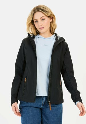 Zwarte waterdichte jas met capuchon, voorzien van ritsen en leren accenten. Draagt over een lichtblauwe hoodie en gecombineerd met blauwe jeans.