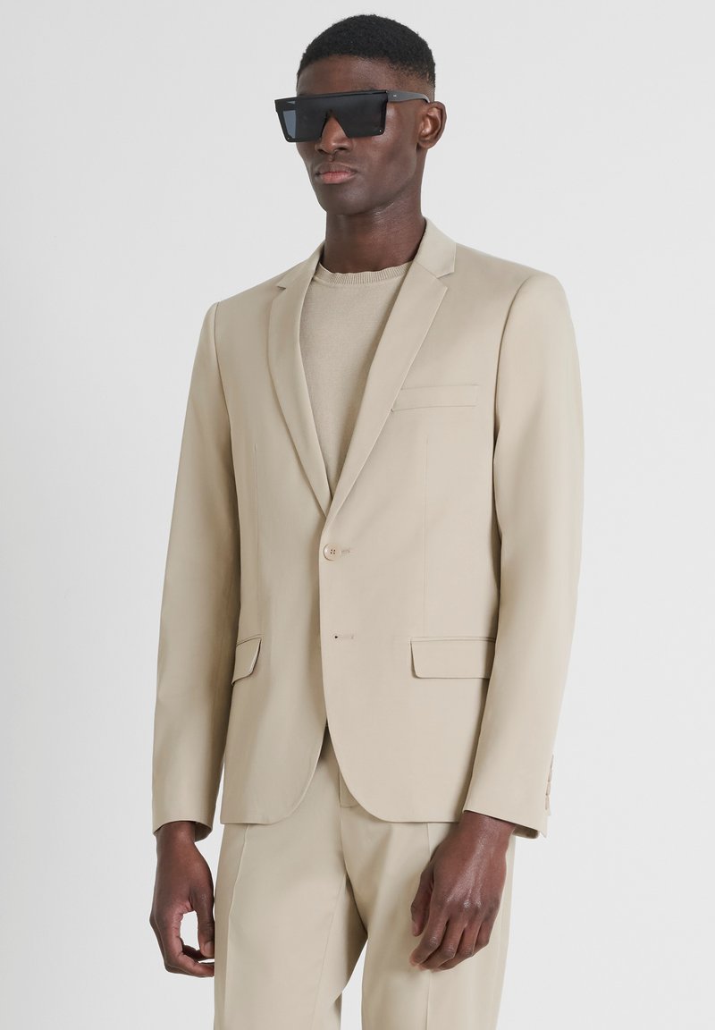 Blazer beige con rever a punta, chiusura a un bottone e due tasche frontali. Abbinato a un maglione beige chiaro e occhiali da sole neri a forma quadrata.
