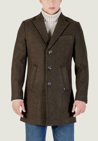 Cappotto di lana marrone con rever a punta, tre bottoni, tasche laterali e una sottile trama, indossato sopra un maglione girocollo crema.