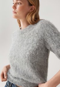 Jasnoszary, puszysty sweter z okrągłym dekoltem i krótkimi rękawami. Miękka struktura, lekko luźny krój, z prążkowanym dołem i mankietami.