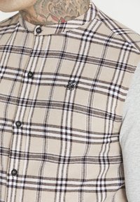 Ruitenhemd met beige, bruin en wit ruitpatroon; voorzien van zwarte knopen en aan één zijde grijze, lange mouwen.