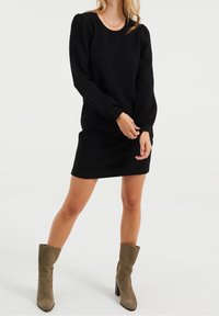 Svart långärmad klänning med en texturerad tyg, rund halsringning och figursydd siluett, tillsammans med olivgröna knähöga stövlar.