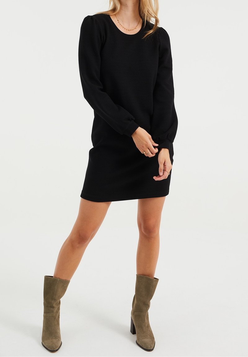 Svart långärmad klänning med en texturerad tyg, rund halsringning och figursydd siluett, tillsammans med olivgröna knähöga stövlar.