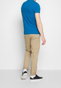 Blaues Baumwoll-T-Shirt mit kurzen Ärmeln, kombiniert mit hellbeigen Chinos. Schwarze Sneakers und weiße Socken sind an den Knöcheln sichtbar.
