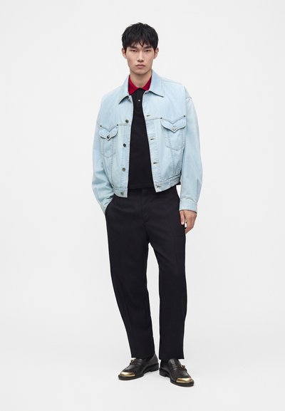 Jeune homme portant une veste en jean bleu clair, un polo noir avec col rouge, un pantalon noir et des chaussures noires avec embouts dorés, debout devant un fond blanc.