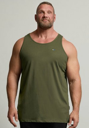 Muskulöser Mann mit Bart blickt nach vorne, trägt ein olivgrünes Tanktop und schwarze Hosen vor einem schlichten, hellen Hintergrund.