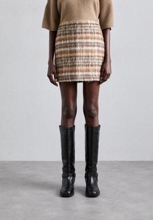 Personne portant un haut beige à manches courtes, une jupe à carreaux beige et marron, et des bottes en cuir noires montant jusqu'aux genoux, debout sur un sol gris.