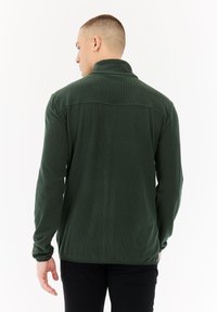 Man met kort haar, met het gezicht van de camera af, in een donkergroene fleecejack met lange mouwen en een zwarte broek, tegen een eenvoudige witte achtergrond.