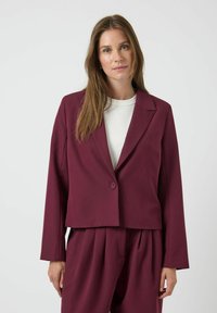 Blazer recortado burdeos con cierre de un solo botón, solapas de muesca y mangas largas. Usado sobre una camisa blanca, que presenta una textura suave.