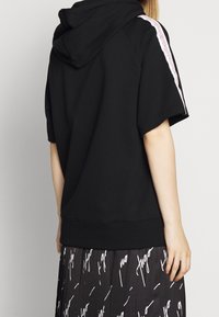 Person iført sort kortærmet hættetrøje med hvid stribe på ærmet og sort nederdel med hvid skrift, set bagfra.