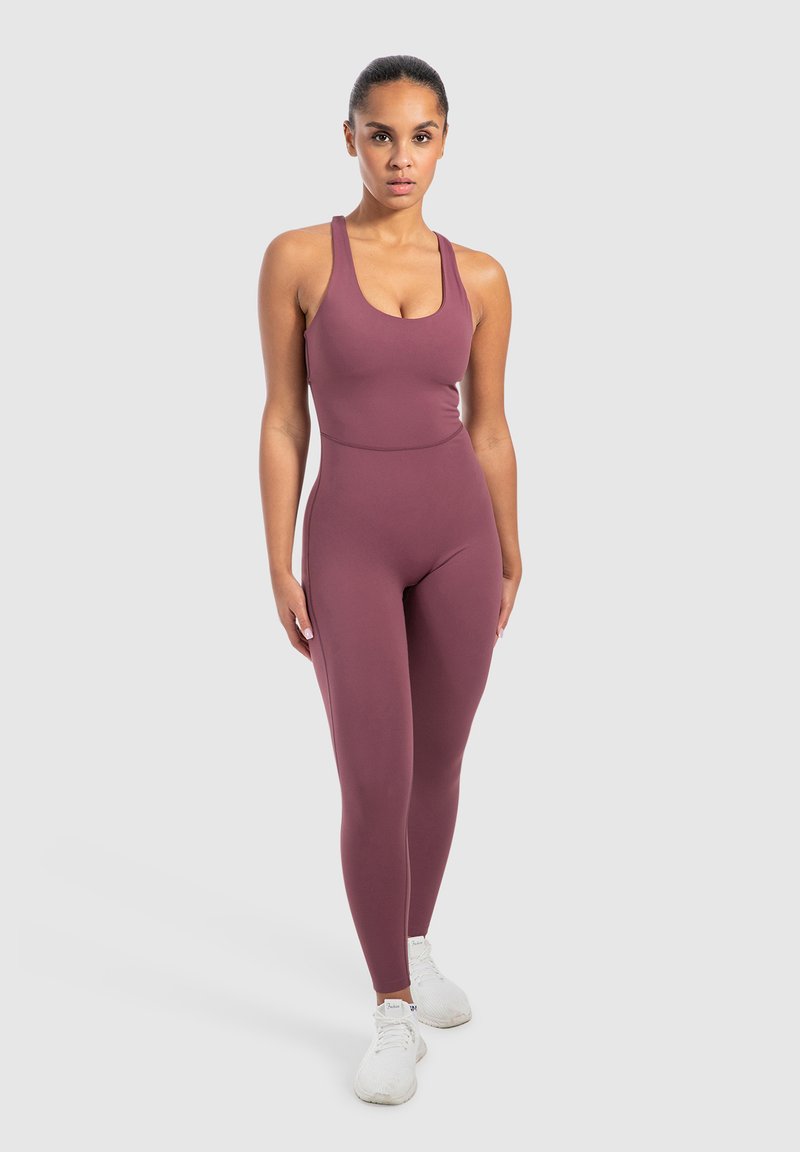 Vrouw staat in een mauve trainingsjumpsuit en witte sneakers tegen een effen lichte achtergrond, recht vooruit kijkend met de handen langs het lichaam.