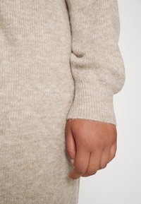 Tessuto beige a coste di maglione con una texture morbida. Primo piano di una mano appoggiata sul materiale, che mostra il dettaglio del polsino.
