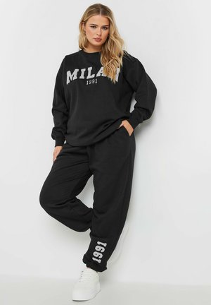 Sweatshirt en coton noir avec "MILAN 1991" en lettres blanches. Associé à un pantalon noir avec imprimé "1991", porté avec des baskets blanches.