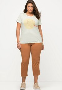 T-shirt in cotone verde chiaro con design grafico astratto; abbinato a pantaloni corti marroni e sneakers beige. Tessuto morbido, taglio rilassato.