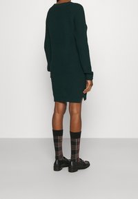 Abito lavorato a maglia verde scuro con maniche lunghe, spacchi laterali e texture a coste. Abbinato a scarpe nere e calze a quadri.