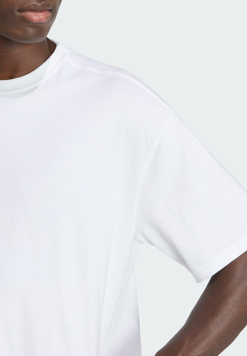 Overkrop af person iført almindelig hvid kortærmet T-shirt mod lysegrå baggrund, med afslappet kropsholdning.