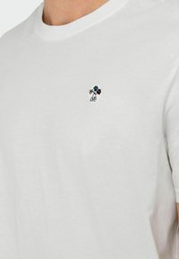 White cotton t-shirt with a round neckline and short sleeves, featuring small multicolored floral embroidery on the left chest area.