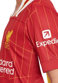 Maglietta rossa con motivo a griglia gialla, con logo I.F.C. e branding "Expedia". Collo bianco, tessuto testurizzato, maniche corte.