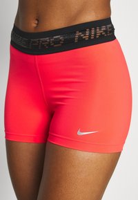 Persona que lleva puestos pantalones cortos Nike Pro de color coral brillante con una cinturilla negra y el logo reflectante en blanco de Nike en el muslo izquierdo.