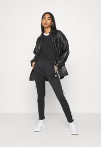 Camisa de imitação de pele preta, t-shirt preta e calças de treino pretas com riscas brancas laterais, combinadas com ténis brancos. Um conjunto casual que se destaca.