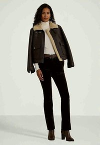 Moteris vilki tamsiai rudą vilnos striukę, užsidėjusi baltą golfo megztinį, juodas kelnes ir rudus aukštakulnius batus, stovinti prieš šviesų foną.