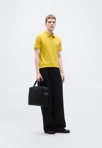 Polo jaune à manches courtes, associé à un pantalon noir ample et tenant un sac noir. Présente un logo discret sur le polo.