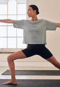 Naine hallis särgis ja mustades lühikestes pükstes teeb heledas toas suure aknaga mustal joogamatil sõdalase joogaasendit.