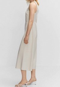 Robe sans manches beige avec une coupe décontractée, longueur genoux, dotée d'une texture lisse et de détails de couture sur les côtés. Portée avec des sandales blanches à lanières.