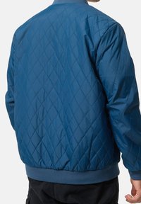 Man draagt een blauwe doorgestikte jas met diamantpatroon, geribde zoom en manchetten, gecombineerd met een zwarte broek, kijkt weg van de camera.